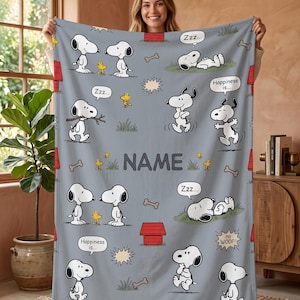 May include: A light gray fleece blanket featuring a repeating pattern of Snoopy and Woodstock in various poses, red doghouses, bones, and speech bubbles with text including "Happiness is..." and "no WOOF!" with a central area for a custom name.