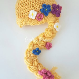 Könnte beinhalten: Ein gelber gehäkelter Babyhut mit einer langen, geflochtenen Garnverlängerung, die einem Zopf ähnelt. Der Hut ist mit kleinen, bunten gehäkelten Blumen in Weiß, Rosa, Blau und Lila verziert. Der Text "Craftsy Momma" ist sichtbar.