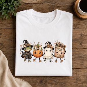 Puede incluir: Una camiseta blanca con un gráfico de una rana con sombrero de bruja, una calabaza sonriente, un fantasma con sombrero de bruja y un cartel que dice SPOOKY CUTE. Los personajes están decorados con detalles florales y lazos de leopardo.