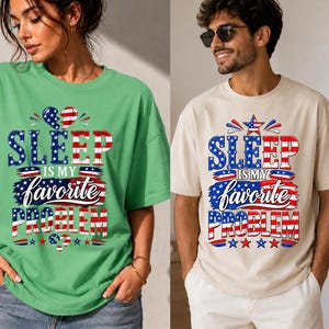 May include: A green t-shirt and a beige t-shirt, each featuring a graphic print with the text 'SLEEP IS MY favorite PROBLEM' in red, white, and blue colors, accented with stars, stripes, and heart shapes in a patriotic style design.