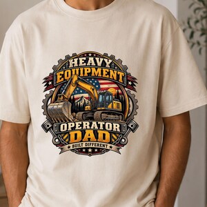 May include: A cream-coloured t-shirt featuring a graphic print of a yellow excavator in front of an American flag and mountain landscape. The text reads HEAVY EQUIPMENT OPERATOR DAD BUILT DIFFERENT, framed by a gear design with two engine pistons.