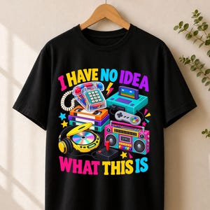 Könnte beinhalten: Ein schwarzes Kurzarm-T-Shirt mit einem bunten Grafikdruck von Retro-Elektronik, darunter ein Festnetztelefon, eine Spielkonsole, VHS-Kassetten, ein CD-Player und ein Ghettoblaster mit dem Text I HAVE NO IDEA WHAT THIS IS in hellen Buchstaben.