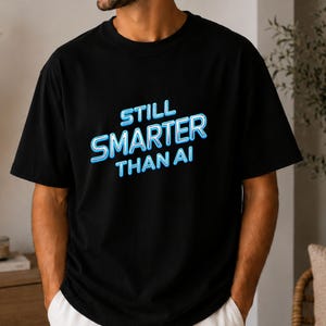Op de afbeelding: Een zwart T-shirt met korte mouwen en de tekst "STILL SMARTER THAN AI" in een lichtblauw neon-stijl lettertype op de borst. Het shirt heeft een ronde hals en een standaard pasvorm, wat zorgt voor een eenvoudige en strakke uitstraling.