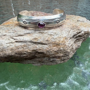 May include: A silver cuff bracelet featuring a central round faceted purple gemstone. The metal band displays engraved geometric patterns, including diamond shapes and linear textures, with an open back design for a slip-on fit.