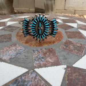 May include: A silver cuff bracelet featuring a central cluster of needle-point turquoise stones arranged in a sunburst pattern. The metalwork includes small silver beads between the elongated blue stones, creating a symmetrical design on a band.