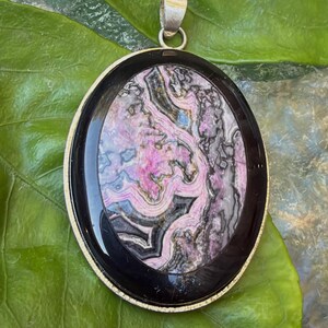May include: An oval pendant featuring a polished rhodochrosite stone with intricate pink, gray, and black banded patterns, encased in a smooth black frame with a silver-toned metal bail at the top for attachment to a necklace chain.
