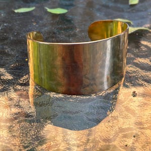 Puede incluir: Una pulsera tipo brazalete ancha de metal dorado pulido con una superficie lisa y reflectante y un diseño abierto en forma de C. La pulsera presenta un ancho constante en toda su banda, mostrando un acabado metálico que refleja la luz.
