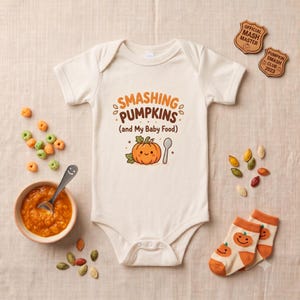 May include: A cream-colored short-sleeve baby bodysuit featuring the text SMASHING PUMPKINS (and My Baby Food) in orange and brown, with a cartoon pumpkin and spoon graphic. The bodysuit is surrounded by small orange cereal pieces and socks.