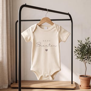 May include: A cream-coloured short-sleeve baby bodysuit featuring the text "BABY" in a simple sans-serif font above the name "Braxton" in a cursive script, with a small grey heart icon centred below the name, all printed on the front.