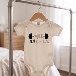 Peut inclure: Un body pour bébé à manches courtes de couleur crème avec une impression graphique d'une barre de musculation noire et le texte MILK, THEN BURPEES. Le mot MILK est en rose et bleu sourds, et le mot BURPEES est en lettres bleues et roses.
