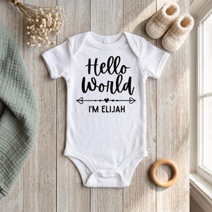Peut inclure: Un body pour bébé blanc à manches courtes avec le texte noir Hello World et I'M ELIJAH accompagné d'un motif de flèche et de cœur au centre. Le vêtement est présenté avec une paire de petits chaussons en tricot de couleur crème.