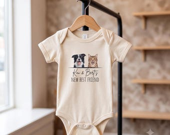 May include: A cream-coloured short-sleeve baby bodysuit featuring a graphic of a black and white dog and a brown tabby cat. Below the animals, the text reads "Kai & Bert's NEW BEST FRIEND" in black lettering. The garment hangs on a wooden hanger.