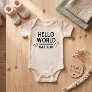 Peut inclure: Un body pour bébé à manches courtes de couleur crème avec le texte imprimé en noir HELLO WORLD I'M ELIJAH accompagné de deux petites icônes d'empreintes de pieds. Le vêtement présente une encolure ronde et des boutons-pression à l'entrejambe.