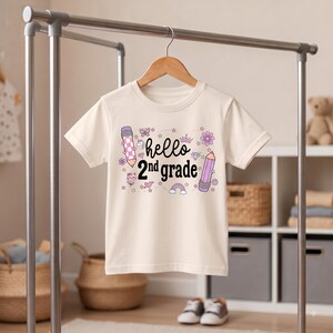 Può includere: Una t-shirt a maniche corte color crema con la scritta nera "hello 2nd grade" stampata sul petto. Il design include illustrazioni di due matite, una corona, un arcobaleno, una farfalla, un fiore, un diamante e un piccolo muso di gatto.