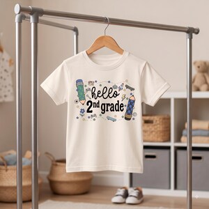 Può includere: Una t-shirt a maniche corte color crema con una stampa grafica che riporta il testo "hello 2nd grade" in nero. Il design include illustrazioni di due matite, un razzo, uno skateboard, un teschio di dinosauro, un controller di gioco e stelle.