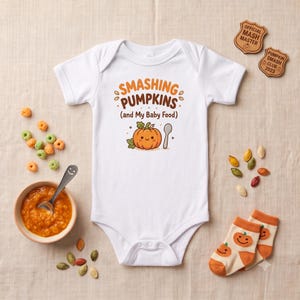 May include: A white short-sleeved baby bodysuit featuring a graphic of a smiling orange pumpkin and a spoon. The text reads SMASHING PUMPKINS (and My Baby Food) in brown and orange. The design includes small orange leaf accents and dots.
