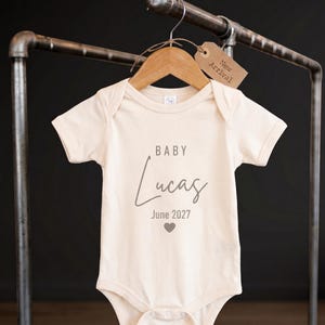 May include: A cream-coloured baby bodysuit featuring the printed text BABY Lucas and June 2027 with a small heart icon. The garment is displayed on a wooden hanger with a brown paper tag attached that reads New Arrival in black ink.