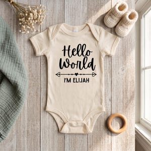 Peut inclure: Un body pour bébé à manches courtes de couleur crème avec le texte noir Hello World et I'M ELIJAH accompagné d'une flèche décorative et d'un cœur. Le vêtement est présenté avec une paire de petits chaussons tricotés et un anneau en bois.