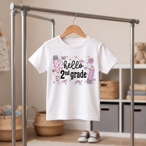 Può includere: Una t-shirt bianca a maniche corte con il testo nero "hello 2nd grade" circondato da illustrazioni colorate di matite, una farfalla, una corona, diamanti, un gatto, un cuore, un arcobaleno e fiori nelle tonalità del rosa e del viola.