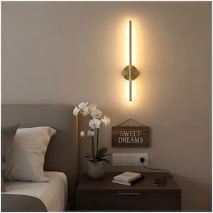 Puede incluir: Un aplique de pared vertical y cilíndrico con acabado en oro metálico que emite un brillo cálido y suave. La lámpara está montada en la pared sobre una mesita de noche, con una base circular central y una varilla larga y delgada.