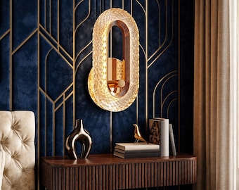 Könnte beinhalten: Eine ovale Wandleuchte mit einem texturierten, klaren kristallinen Außenrahmen und einer goldfarbenen Innenstruktur. Die Leuchte ist an einer dunkelblauen Wand mit goldenen geometrischen Linien über einem schwebenden Konsolentisch aus Holz montiert.