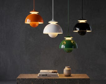 Könnte beinhalten: Vier kuppelförmige Pendelleuchten in Orange, Weiß, Grün und Schwarz hängen von der Decke. Jede Lampe hat eine kleinere Kuppel unter einer größeren und strahlt ein sanftes Licht aus. Darunter liegt ein Bücherstapel auf einem gesprenkelten Block.