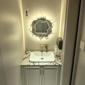 Water splash mirror wall light installed above a modern bathroom vanity, combining a reflective acrylic mirror with decorative integrated LED lighting.