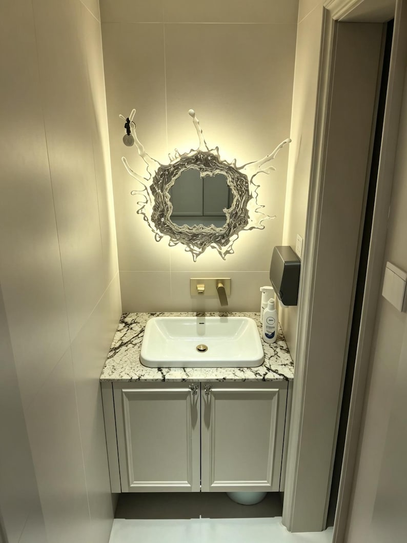 Water splash mirror wall light installed above a modern bathroom vanity, combining a reflective acrylic mirror with decorative integrated LED lighting.