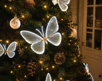 Könnte beinhalten: Mehrere beleuchtete Schmetterlingsornamente mit durchscheinenden weißen Flügeln und leuchtenden Rändern, die an einem grünen Weihnachtsbaum befestigt sind. Die Schmetterlinge haben zarte Flügelmuster und sind zwischen verschiedenen goldenen und weißen Kugeln platziert.