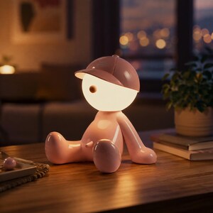 Op de afbeelding: Een tafellamp in de vorm van een zittend figuur met een glad, lichtroze lichaam en een bijpassende roze honkbalpet. Het hoofd is een gloeiende, bolvormige witte lamp die een zacht, warm licht uitstraalt voor een aangename sfeer.