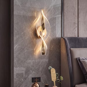 May include: A wall-mounted light fitting featuring a central polished gold-tone vertical bar with two spiralling, translucent acrylic wings that emit a warm glow. The design includes a circular base plate and integrated LED lighting elements.