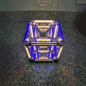 Könnte beinhalten: Eine quadratische Unendlichkeitsspiegel-Würfellampe mit einem klaren Acrylrahmen und interner LED-Beleuchtung. Das Gerät zeigt einen sich wiederholenden Tunneleffekt aus leuchtend warmweißen horizontalen Linien mit blauen Akzenten.