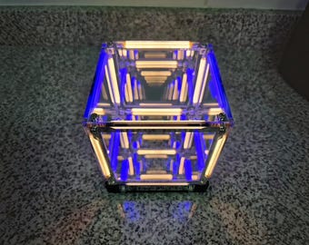 Könnte beinhalten: Eine quadratische Unendlichkeitsspiegel-Würfellampe mit einem klaren Acrylrahmen und interner LED-Beleuchtung. Das Gerät zeigt einen sich wiederholenden Tunneleffekt aus leuchtend warmweißen horizontalen Linien mit blauen Akzenten.