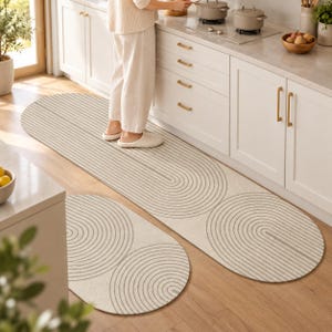 Puede incluir: Un juego de dos alfombras de cocina ovaladas con una base de color beige y un patrón repetitivo de finas líneas y semicírculos concéntricos de color gris oscuro. Las alfombras están colocadas sobre un suelo de madera clara en una cocina.