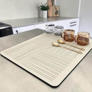 May include: A rectangular off-white placemat featuring a pattern of thin, dark grey horizontal and vertical lines. Two clear textured glass tumblers, two amber-colored glass mugs, and two gold-toned metal spoons are arranged on the surface.