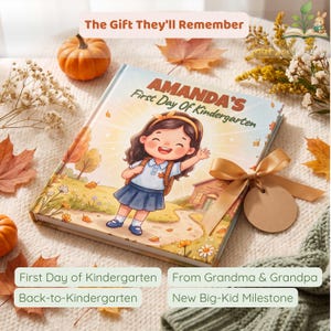 May include: A hardcover children's book titled Amanda's First Day Of Kindergarten featuring an illustration of a child with a backpack in front of a school building, decorated with a gold ribbon and a blank circular tag on the cover.