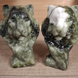 May include: Two carved stone lion head sculptures. The left lion is dark green with black veining. The right lion features a mix of dark green and white stone. Both sculptures show detailed manes and facial features in a vertical bust orientation.