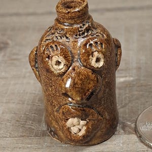 May include: A small brown ceramic face jug with a glossy finish, featuring protruding eyes, a prominent nose, and an open mouth revealing small teeth. The jug has a handle at the top and is positioned next to a US quarter coin for scale reference.