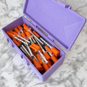 Op de afbeelding: Een lichtpaarse plastic opbergdoos gevuld met meerdere doorzichtige plastic injectiespuiten met feloranje doppen en plunjers. Elke spuit heeft zwarte maatstreepjes op de cilinder en de doos is voorzien van een scharnierend deksel.