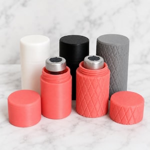 May include: A collection of cylindrical containers with removable caps. Two containers are coral pink with a diamond-textured pattern, while three others are smooth in white, black, and gray. Each container holds a small silver metal vial inside.