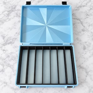 Peut inclure: Un boîtier de rangement rectangulaire bleu clair avec un couvercle à charnière présentant un motif géométrique en rayons de soleil. L'intérieur comprend un insert noir avec huit fentes verticales, maintenu par des loquets noirs aux extrémités.