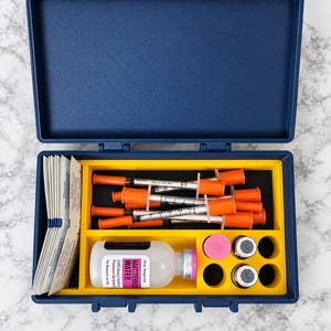 May include: A navy blue plastic storage case with yellow latches containing medical supplies. Inside are several orange-capped syringes, a 30 mL bottle labeled BACTERIOSTATIC WATER, two small metal vials, a pink cap, and white packets.