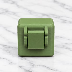 May include: A small, matte olive green square storage box with a hinged lid and a central latch mechanism on the front. The container features a smooth, uniform finish and a compact, cubic shape with rounded edges, designed for secure closure.