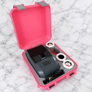 Puede incluir: Un maletín rígido de color rosa brillante con cierres grises, que contiene una impresora de etiquetas portátil de color gris oscuro con la marca NIIMBOT impresa en el frente, tres rollos de etiquetas circulares y un cable USB negro en espuma.