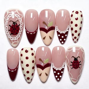 Könnte beinhalten: Ein Set aus zehn mandelförmigen Press-on-Nägeln mit verschiedenen Designs, darunter rote Marienkäfer in weißen Spitzenrahmen, dunkelrote French-Tips mit weißen Nähten, stilisierte Apfelscheiben und cremefarbene Nägel mit roten Punkten.