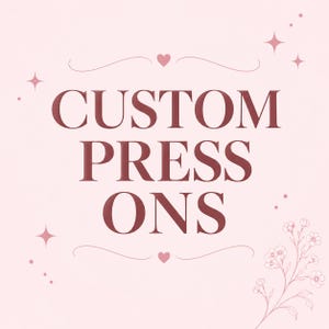 May include: A light pink graphic featuring the dark maroon text CUSTOM PRESS ONS in a serif font. The design includes decorative flourishes, small heart icons above and below the text, four-pointed star accents, and a floral line drawing.