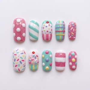 May include: A set of ten press-on nails featuring various colorful designs including pink with white polka dots, teal and white stripes, cupcake motifs, plaid patterns, multi-colored sprinkles, and raised circular accents in a glossy finish.