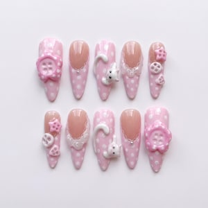 Può includere: Un set di dieci unghie finte a mandorla con base rosa chiaro e pois bianchi. I design includono decorazioni 3D come fiori rosa, piccole figure di gatti bianchi, ciondoli a forma di stella, dettagli a bottone e bordi in pizzo bianco.