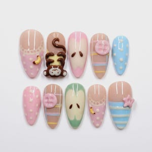 May include: A set of ten almond-shaped press-on nails featuring various designs including a 3D brown monkey, pink and blue polka dots, plaid patterns, apple slice motifs, a pink button, and a small pink star accent on a glossy finish.