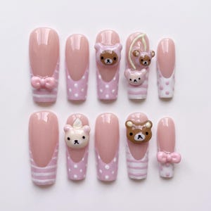 Puede incluir: Un juego de diez uñas postizas con una paleta de colores rosa y blanco. Los diseños incluyen puntas francesas, lunares blancos, rayas horizontales, dijes de osos en tres dimensiones y pequeños lazos rosas con acabado brillante.